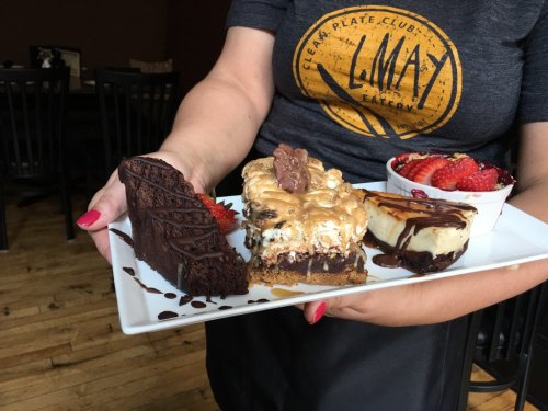 A person holds a plate with three desserts: chocolate cake, a layered treat, and a cheesecake topped with strawberries.