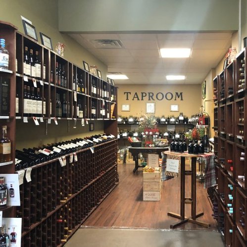 Interior of a wine taproom with wooden shelves filled with bottles, a table, and decorative plants.