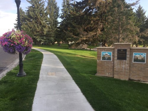 A paved walkway lined with trees and flowers, leading to a brick monument with plaques in a green park setting.