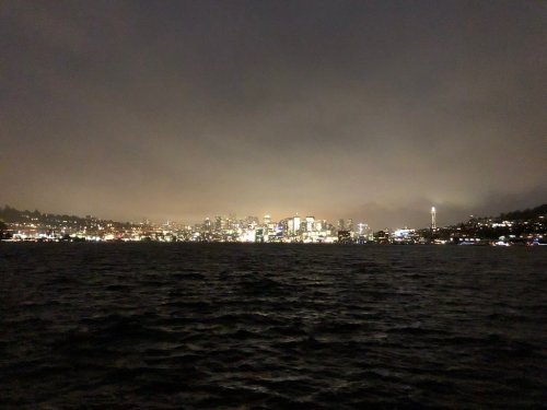 City skyline at night with lights reflecting on water, shrouded in fog, featuring a tall tower in the distance.