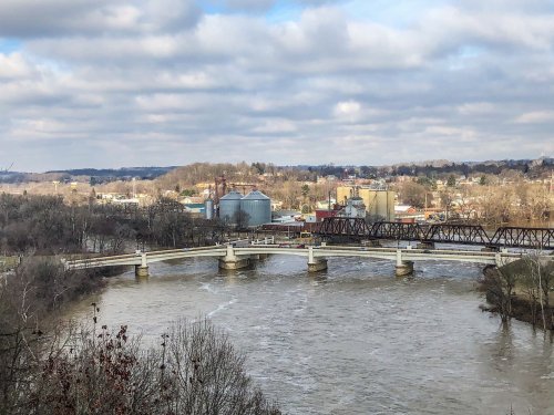 A river flows under a bridge, with industrial buildings and trees lining the banks against a cloudy sky.