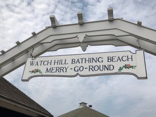 Sign for Watch Hill Bathing Beach and Merry-Go-Round, with decorative elements, under a cloudy sky.
