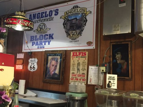 A restaurant wall featuring a banner for Angelo's 50th anniversary, Elvis posters, and Route 66 signage.