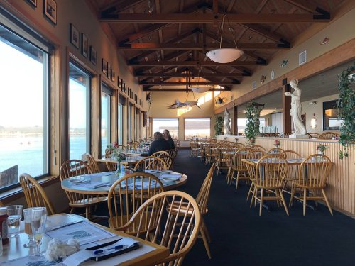 Interior of a restaurant with wooden chairs, large windows overlooking water, and a cozy atmosphere.