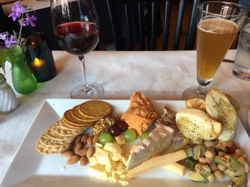 A platter of cheese, crackers, nuts, and spreads, accompanied by a glass of red wine and a beer.