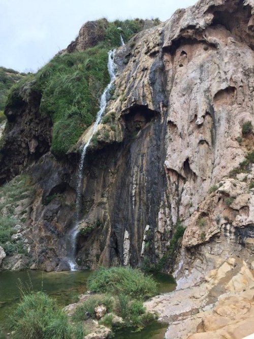 A waterfall cascading down rocky cliffs, surrounded by lush greenery and a small pool at the base.