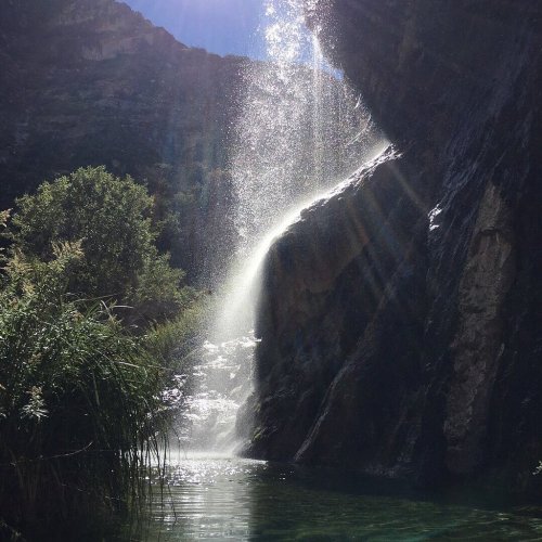 A waterfall cascades down rocky cliffs into a serene pool, surrounded by lush greenery and sunlight filtering through.