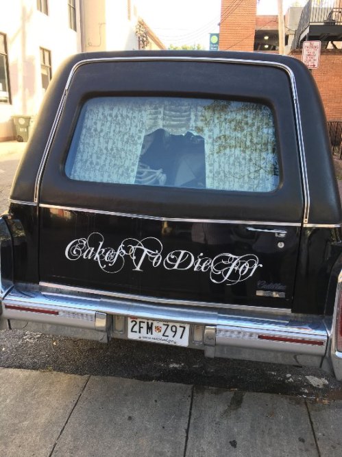 A black hearse with a decorative rear window and the phrase "Cakes to Die For" written on it.