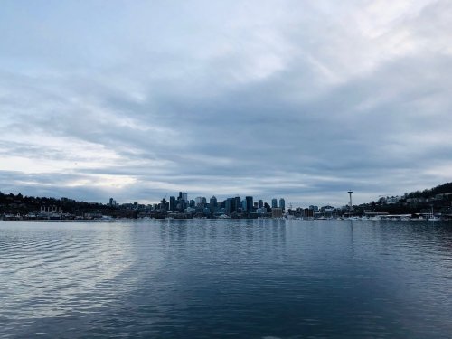 A calm body of water reflects a cloudy sky and the skyline of a city with tall buildings in the background.
