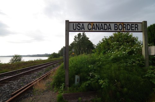 Sign marking the USA-Canada border, with train tracks and lush greenery in the background under a cloudy sky.