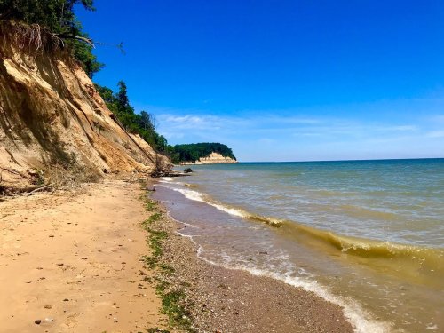 A sandy beach with gentle waves, cliffs in the background, and a clear blue sky above.
