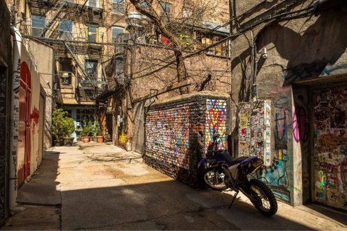 Narrow alleyway with graffiti-covered walls, a motorcycle parked, and greenery peeking through in an urban setting.