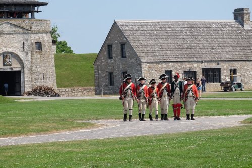 A group of soldiers in historical uniforms march near a stone fort and buildings on a sunny day.