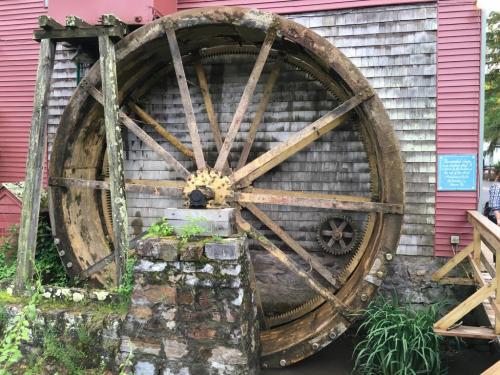 A large, weathered water wheel stands against a wooden building, surrounded by greenery and stone.
