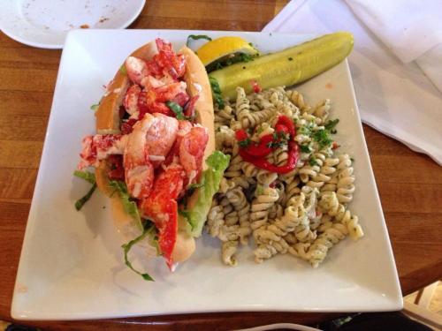 A lobster roll with lettuce, a pickle, and a side of pesto pasta on a white plate.