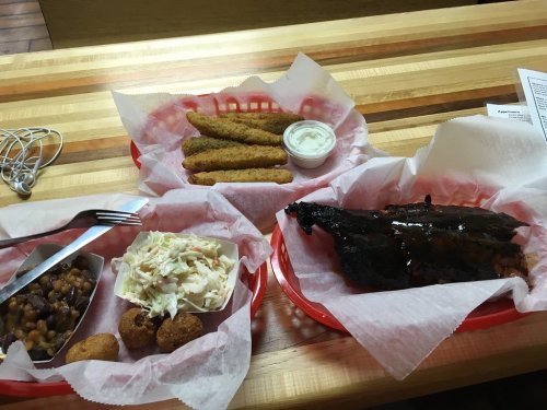 A wooden table with two red baskets containing ribs, coleslaw, cornbread, and fried pickles with dipping sauce.