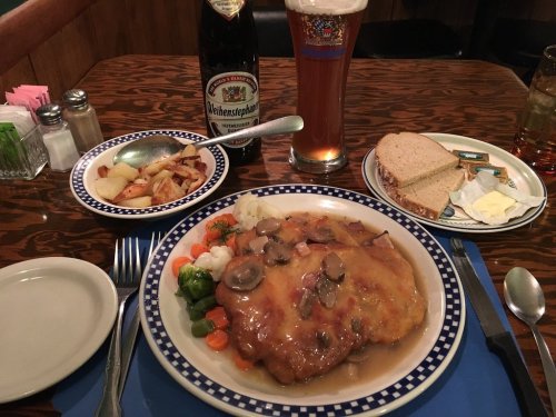 A plate of chicken with mushroom sauce, mixed vegetables, fries, bread, and a beer on a wooden table.
