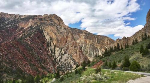 A scenic view of rugged mountains with colorful rock formations under a partly cloudy sky. Green vegetation in the foreground.