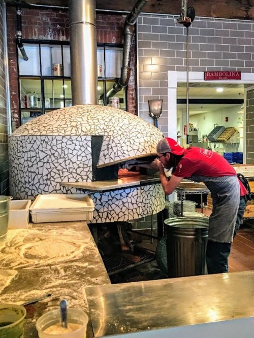 A chef in a red shirt places a pizza into a large, mosaic-tiled wood-fired oven in a bustling kitchen.