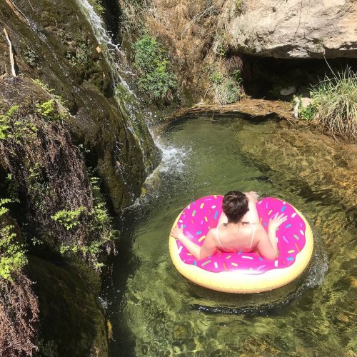 A person relaxes on a pink donut-shaped float in a serene, green pool near a small waterfall.