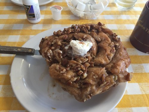 A large, cinnamon roll topped with pecans and whipped cream on a white plate, set on a yellow checkered tablecloth.