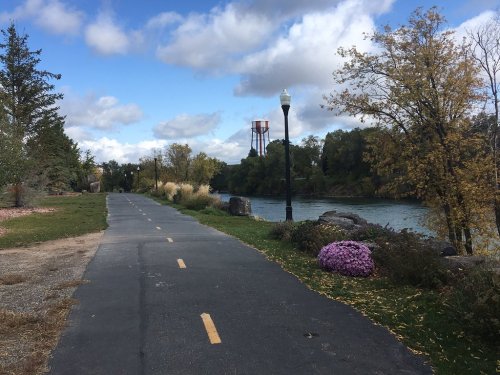 A scenic pathway along a river, lined with trees and flowers, under a partly cloudy sky. A water tower is visible in the distance.