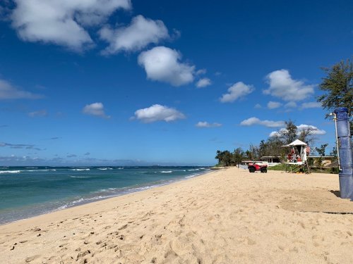 A sandy beach with gentle waves, blue sky, and a lifeguard tower in the distance under fluffy white clouds.