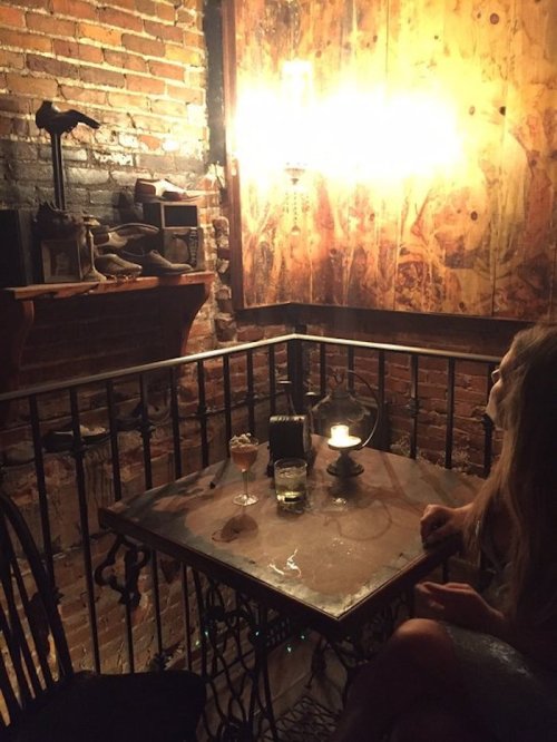 A cozy, dimly lit corner of a rustic bar with a small table, drinks, and exposed brick walls.