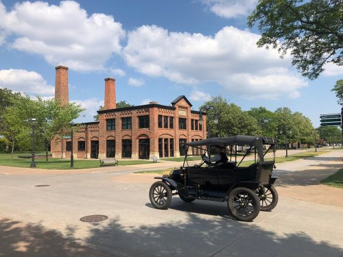 A vintage black car drives past a historic brick building with smokestacks under a blue sky with fluffy clouds.