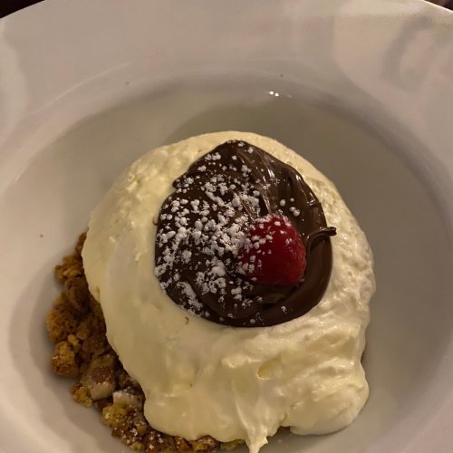 A creamy dessert topped with chocolate, a raspberry, and powdered sugar, served on a bed of crushed cookies.