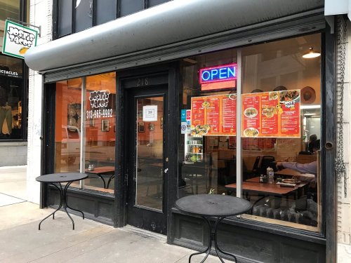 Exterior of a restaurant with an "OPEN" sign, featuring menu posters on the window and outdoor tables.