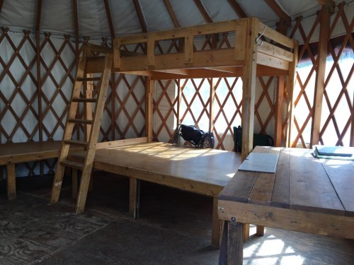 Interior of a yurt featuring a wooden loft bed, ladder, and simple wooden furniture. Natural light filters in.