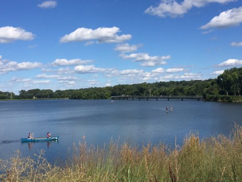 A serene lake scene with two people kayaking and others paddleboarding, surrounded by lush greenery and fluffy clouds.