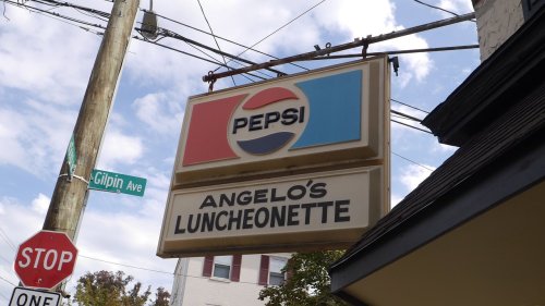 Sign for Angelo's Luncheonette featuring the Pepsi logo, with a stop sign and street sign in the background.