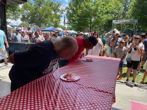 Two competitors lean over a table, eating from plates during a food contest, with a crowd watching in the background.