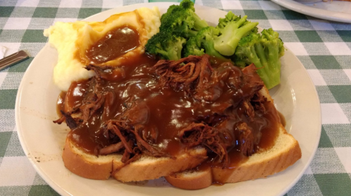 A plate of shredded meat in gravy on bread, served with mashed potatoes and broccoli on a checkered tablecloth.
