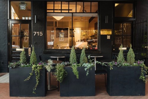 A cozy café exterior with potted plants and small tables outside, featuring large windows and a welcoming atmosphere.