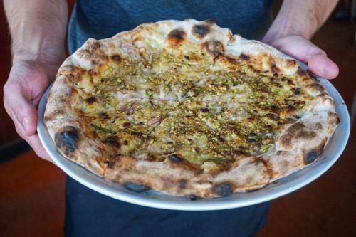 A person holds a freshly baked pizza topped with herbs and crushed nuts on a white plate.