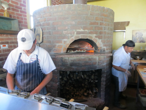 Two chefs work in a rustic kitchen with a large brick oven, preparing food near a stack of firewood.