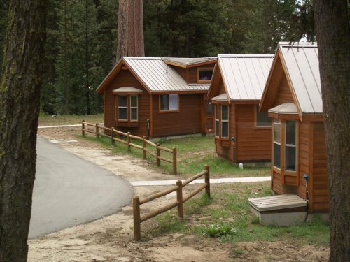 Three small wooden cabins with metal roofs, surrounded by trees and a gravel path, in a serene outdoor setting.