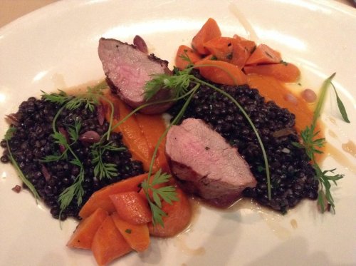 A plate featuring sliced meat, black lentils, and orange carrots, garnished with fresh herbs.