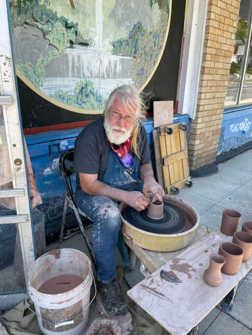 An elderly man with long white hair and a beard shapes clay on a pottery wheel outside a shop.
