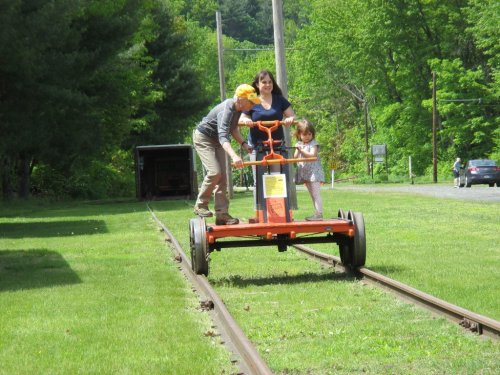 A man, woman, and child ride on a handcar along a grassy railway track surrounded by trees.