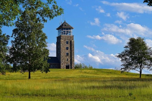 A stone tower stands on a grassy hill under a blue sky with scattered clouds, surrounded by trees.