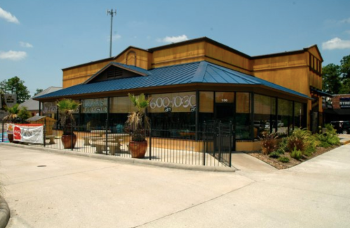 A yellow building with a blue roof and large windows, featuring outdoor seating and palm plants.
