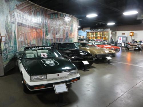 A display of classic cars in a museum, featuring an Oldsmobile and various vintage models against a mural backdrop.