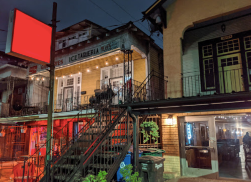 A colorful building at night with a red sign, featuring a taqueria and outdoor seating under string lights.