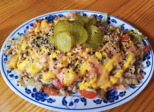 A colorful plate of rice topped with ground beef, tomatoes, pickles, and drizzled with mustard and a creamy sauce.