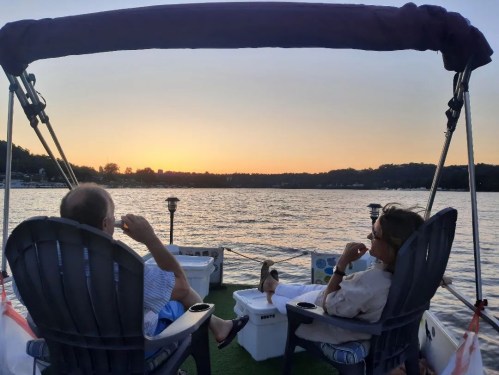 A couple relaxes on a boat at sunset, enjoying the view over the calm water. A small dog sits nearby.