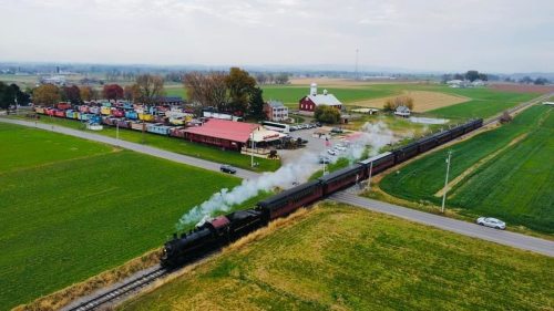 A vintage steam train travels through a rural landscape with fields, a farm, and colorful buildings in the background.
